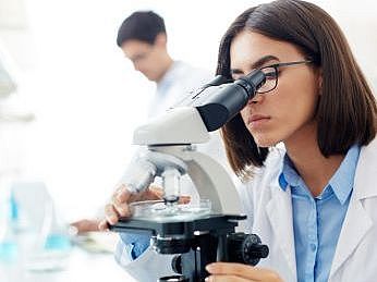 Eine Frau mit weißem Kittel und Brille schaut durch ein Mikroskop. Im Hintergrund arbeitet ein Mann