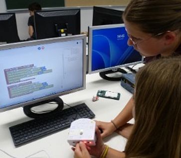 Zwei Schülerinnen programmieren an einem Computerbildschirm einen weißen Thymio-Roboter.