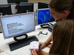 Zwei Schülerinnen programmieren an einem Computerbildschirm einen weißen Thymio-Roboter.