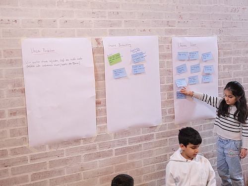 Auf diesem Bild sieht man Kinder an großen Plakaten, die an der Wand kleben. Sie finden gemeinsam Ideen für ihren Prototypen und diese Ideen werden mit Post it´s auf das Plakat geklebt.