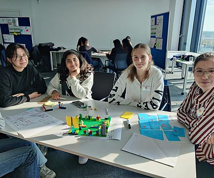 Vier Mädchen sitzen um einen Tisch auf dem eine LEGO-Landschaft aufgebaut wurde und lächeln in die Kamera.