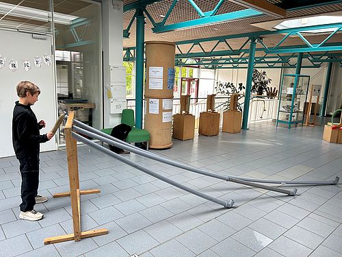 Ein Junge steht in einer hellen Schulaula an einer Experimentierstation der MINIPHÄNOMENTA. Die Station besteht aus einem Holzgestell, an dem mehrere lange, graue Kunststoffrohre schräg nach unten verlaufen. Der Schüler hält ein Objekt an die Rohröffnung, um Akustikphänomene zu erforschen. Im Hintergrund ist der großzügige Flurbereich der Schule mit weiteren Lernstationen zu sehen.