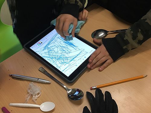 Auf diesem Bild sieht man ein Tablet mit einem Touchscreen, auf dem mit unterschiedlichen Materialien gemalt und geschrieben wird. Es sind Kinderhände mit Stiften, Löffeln aus Metall und Plastik und ein schwarzer Handschuh zu sehen.