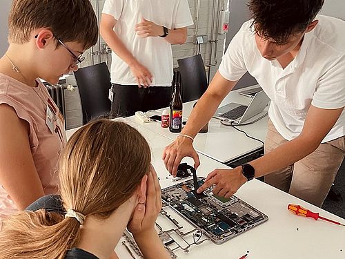 Zwei Jugendliche schauen aufmerksam zu, wie ein Betreuer die Komponenten eines geöffneten Laptops auf dem Tisch erklärt.
