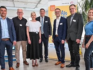 Sieben lächelnde Personen stehen nebeneinander bei einer Auftaktveranstaltung vor einem gelben Werbeaufsteller in einem hellen Büro.