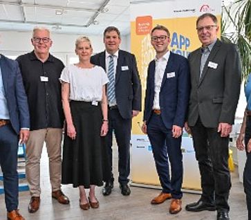 Sieben lächelnde Personen stehen nebeneinander bei einer Auftaktveranstaltung vor einem gelben Werbeaufsteller in einem hellen Büro.