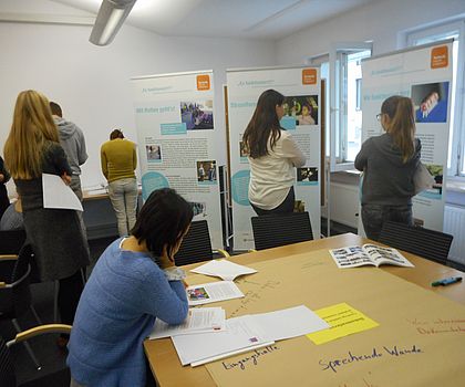 Auf dem Foto sind Teilnehmende der Veranstaltung zu sehen. In einem Seminarraum sind Roll-up´s mit Best-Practice-Projekten von Es funktioniet?! aufgebaut. An einem großen Tisch sitzt eine Teilnehmerin und liest eine Projektdokumentation.
