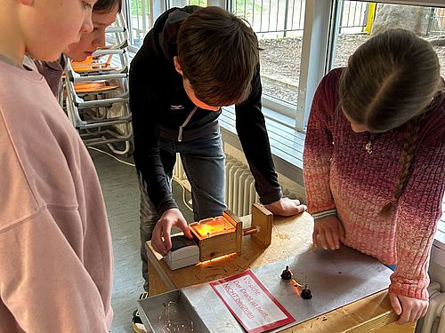 Vier Kinder stehen dicht gedrängt an einer hölzernen Experimentierstation in einem hellen Klassenzimmer vor einem Fenster. Auf dem Tisch der Station befindet sich eine Metallplatte mit einer Warnung, Kabeln und einer kleinen, orange leuchtenden Lichtquelle. Die Kinder blicken aufmerksam und konzentriert nach unten auf das Experiment.