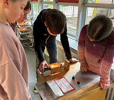 Vier Kinder stehen dicht gedrängt an einer hölzernen Experimentierstation in einem hellen Klassenzimmer vor einem Fenster. Auf dem Tisch der Station befindet sich eine Metallplatte mit einer Warnung, Kabeln und einer kleinen, orange leuchtenden Lichtquelle. Die Kinder blicken aufmerksam und konzentriert nach unten auf das Experiment.