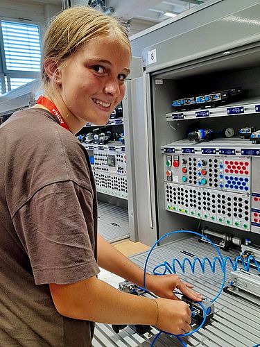 Eine lächelnde Teilnehmerin des Mädchen für Technik-Camps bei ZF Schweinfurt schließt einen blauen Pneumatikschlauch an eine Übungsstation an. Sie schaut direkt in die Kamera.