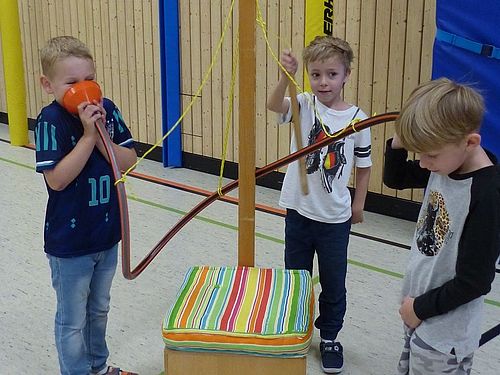 Drei Jungen im Grundschulalter experimentieren in einer Turnhalle an einer MINIPHÄNOMENTA-Station mit einem orangefarbenen Trichter und Schläuchen, um Schallwellen und Töne zu erforschen.