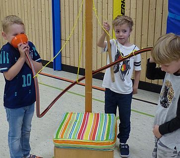 Drei Jungen im Grundschulalter experimentieren in einer Turnhalle an einer MINIPHÄNOMENTA-Station mit einem orangefarbenen Trichter und Schläuchen, um Schallwellen und Töne zu erforschen.
