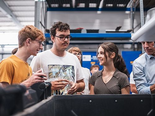 Technik-trifft-Umwelt-Camp bei Kiefel GmbH und TH Rosenheim 1 Mädchen und 2 Jungen unterhalten sich. Im Hintergrund sind Regale zu sehen
