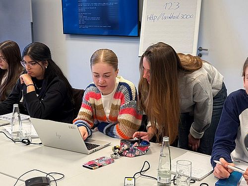 Eine Gruppe von Schülerinnen sitzen an einem Tisch. Sie arbeiten an Laptops. Ein Mädchen beugt sich zu einem anderen Mädchen rüber und erklärt etwas