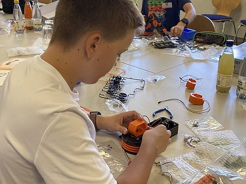 Ein Junge sitzt an eine mTisch . Vor ihm liegen Bauteile des Roboterbausatzes. Er baut 2 Teile zusammen. Im Hintergrund sitzen weitere Jungen