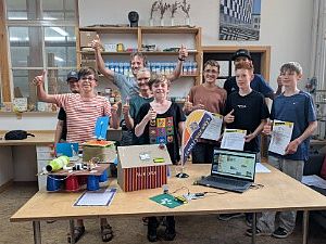 Neun lächelnde Jugendliche und Betreuer der Bildungsinitiative stehen im Werkraum Augsburg hinter einem Tisch und zeigen den Daumen nach oben. Auf dem Tisch sind gebastelte Elektronik-Projekte wie eine Station mit der Aufschrift „Aurora“, ein Laptop mit einem Programmier-Code sowie Urkunden und eine kleine code your way-Fahne zu sehen.