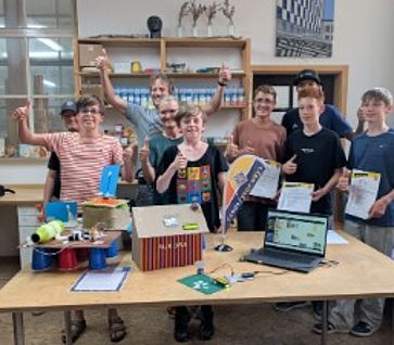 Neun lächelnde Jugendliche und Betreuer der Bildungsinitiative stehen im Werkraum Augsburg hinter einem Tisch und zeigen den Daumen nach oben. Auf dem Tisch sind gebastelte Elektronik-Projekte wie eine Station mit der Aufschrift „Aurora“, ein Laptop mit einem Programmier-Code sowie Urkunden und eine kleine code your way-Fahne zu sehen.