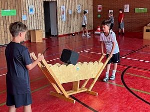 Zwei Grundschulkinder experimentieren in einer Turnhalle gemeinsam an einer hölzernen MINIPHÄNOMENTA-Station mit einem großen, schwarzen Würfel.