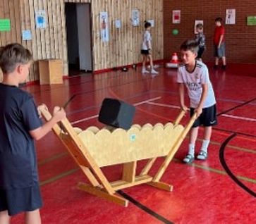 Zwei Grundschulkinder experimentieren in einer Turnhalle gemeinsam an einer hölzernen MINIPHÄNOMENTA-Station mit einem großen, schwarzen Würfel.