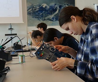 4 Jugendliche sitzen am Tisch. Vor ihnen steht jeweils ein Lötkolben. Ein Mädchen hält ein Sensor-Board in der Hand