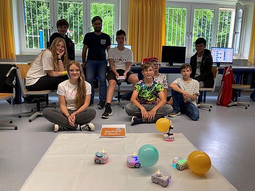 im Vordergrund schreiben 2 Thymio-Radroboter eine 25; Schülerinnen sitzen im Hintergrund