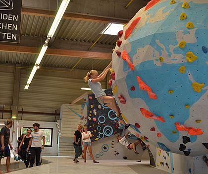 Man sieht eine Boulderhalle. An einer runden Stelle klettert gerade ein Mädchen.