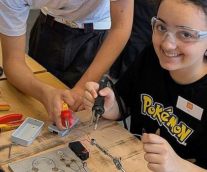 Ein Junge hilft einem sitzenden Mädchen bei  Lötübungen