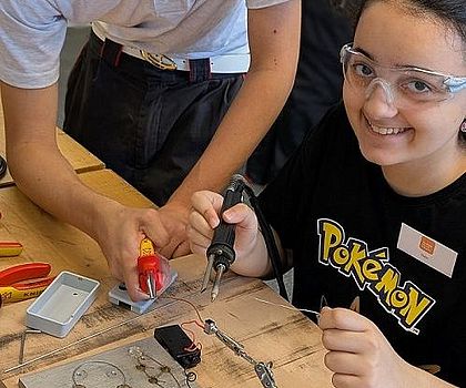 Ein Junge hilft einem sitzenden Mädchen bei  Lötübungen