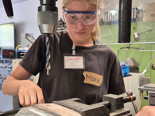 Ein Mädchen mit Schutzbrille konzentriert sich beim Bedienen einer Ständerbohrmaschine in einer Werkstatt.