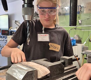 Ein Mädchen mit Schutzbrille konzentriert sich beim Bedienen einer Ständerbohrmaschine in einer Werkstatt.