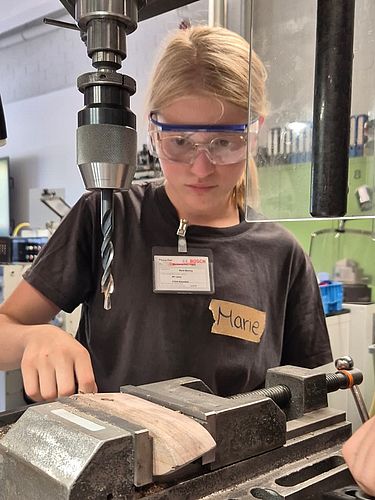 Ein Mädchen mit Schutzbrille konzentriert sich beim Bedienen einer Ständerbohrmaschine in einer Werkstatt.