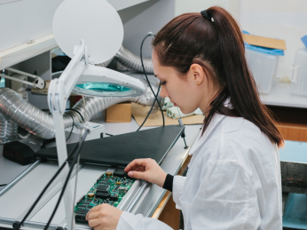 Eine Frau mit braunen Haar und weißem Kittel sitzt an einem Tisch. Vor ihr liegt ein Platine.