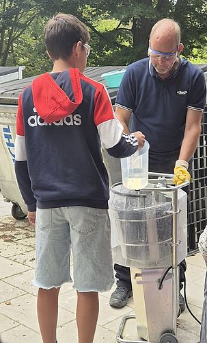 Schüler und Dozent mit Schutzbrillen an einer Betonmischmaschine im Freien, Schüler schüttet Flüssigkeit aus einem Messbecher ein, Workshop zu nachhaltigen Baustoffen.