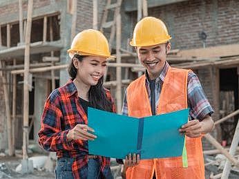 Titelbild Beruf Bauingenieurwesen EIn Mann und eine Frau stehen auf einer Baustelle. Sie tragen einen gelben Baustellenhelm. Sie halten einen Bauplan in der HAnd