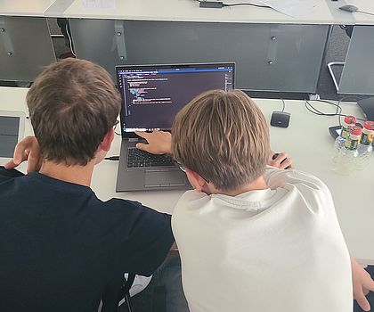 Es sind zwei Jungen von hinten zu sehen, die ihn einen Laptop schauen. 