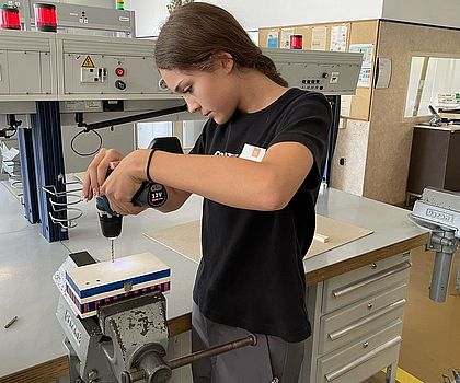 In einem Schrabustock ist eine Holzkiste eingespannt. Ein Mädchen steht davor und bohrt Löcher in die Kiste