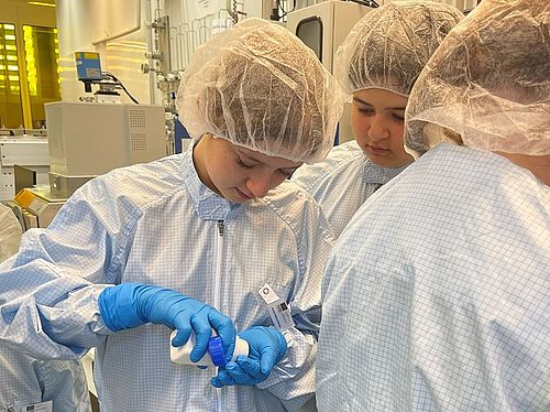Drei Jugendliche in hellblauen Reinraum-Schutanzügen mit Hauben und Handschuhen arbeiten mit einer Dose im Labor.