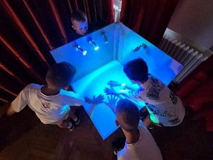 Fünf Kinder betrachten fasziniert ihre Hände in einem weißen Gehäuse, das von innen hellblau beleuchtet wird. Die Kinder stehen dicht gedrängt und blicken von oben in das Gehäuse, während drei Lichter das Innere erhellen.