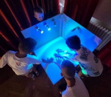 Fünf Kinder betrachten fasziniert ihre Hände in einem weißen Gehäuse, das von innen hellblau beleuchtet wird. Die Kinder stehen dicht gedrängt und blicken von oben in das Gehäuse, während drei Lichter das Innere erhellen.