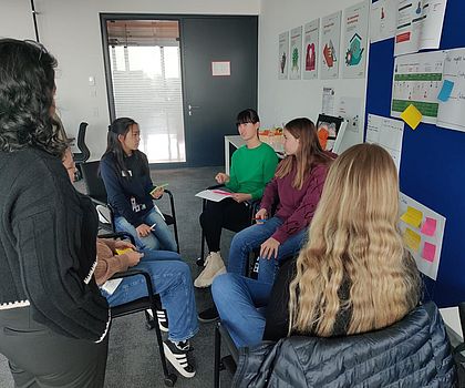 Vier Mädchen sitzen in einem Stuhlkreis vor einer Pinnwand. Ein Mädchen redet mit einer Frau, die man von hinten links im Bild ebenfalls sieht. 