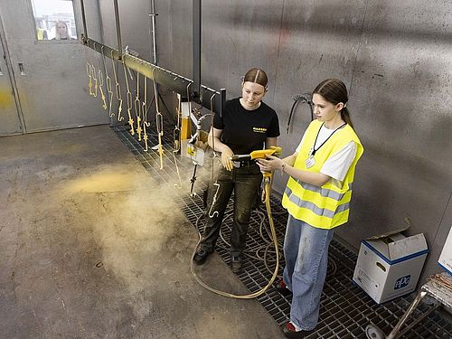 Zwei Mädchen stehen in einer Lackierhalle. Eien hällt eine Lackierpistole in der Hand. Vor ihr sind Metallteile an einer Schnur aufgehangen. Das Mädchen neben ihr erklärt alles
