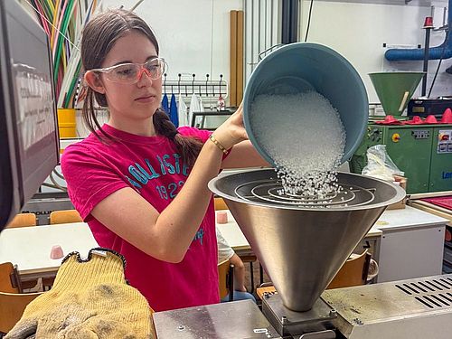 Technik-trifft-Umwelt-Camp bei Kiefel GmbH und TH Rosenheim Ein Mädchen steht vor einem Trichter und füllt Plastikgranulat ein. Sie trägt eine Schuztzbrille