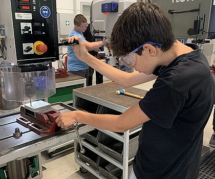 Ein Junge bohrt Löcher in eine Metallplatte. Er steht dafür an der Standbohrmaschine