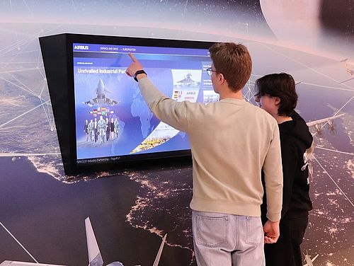Bionik-Camp bei Airbus in Taufkirchen 2 Jugendliche stehen vor einem Bildschirm auf den Flugzeuge von Airbus präsentiert werden