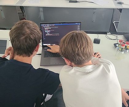 Es sind zwei Jungen von hinten zu sehen, die ihn einen Laptop schauen. 