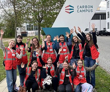 Man sieht 12 Mädchen in roten Warnweste wie sie freudig ihre Arme nach oben strecken. Im Hintergrund ist das Logo der Firma Carbon Campus zu sehen. 