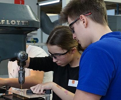 Ein Mädchen bohrt in eine Metalplatte mit einer großen Maschine. Der Ausbilder schaut zu