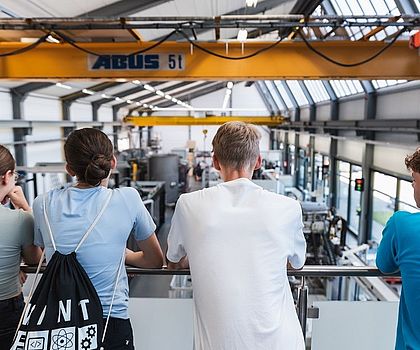 3 Junge und 1 Mädchen schauen von einer Galerie auf die Werkshalle