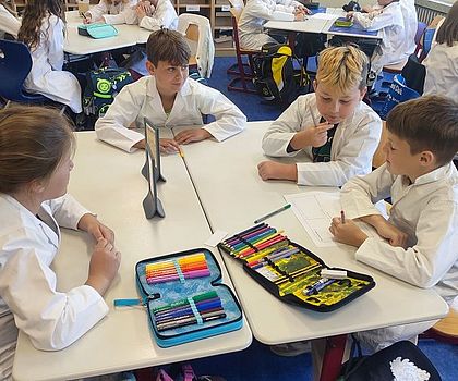 Auf dem Foto sitzen Kinder an Schultischen und teilen und schreiben ihre Vermutungen zur ihrer Forscherfrage auf. 