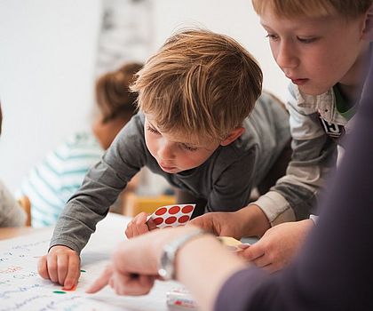 Auf diesem Foto sind Kinder zu sehen, die an einem Tisch sitzen. Auf dem Tisch liegt ein großes, weißes Papier, auf dem die Ideen der Kinder für eine neue Erfindung geschrieben stehen.  Jedes Kind darf mit farbigen Klebepunkten über die Ideen abstimmen. Ein Junge beugt sich gerade über das Papier und klebt seinen Punkt  an eine Idee, die ihm gefällt.
